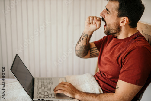 Sleepy man yawling and working on a laptop in the morning from the bed photo