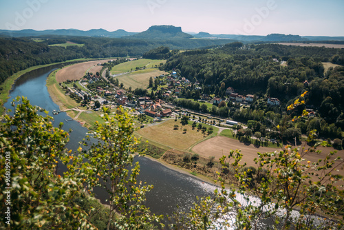 Panoramic view of a picturesque village nestled by a winding river in a green valley with surrounding forests and distant hills under a clear blue sky