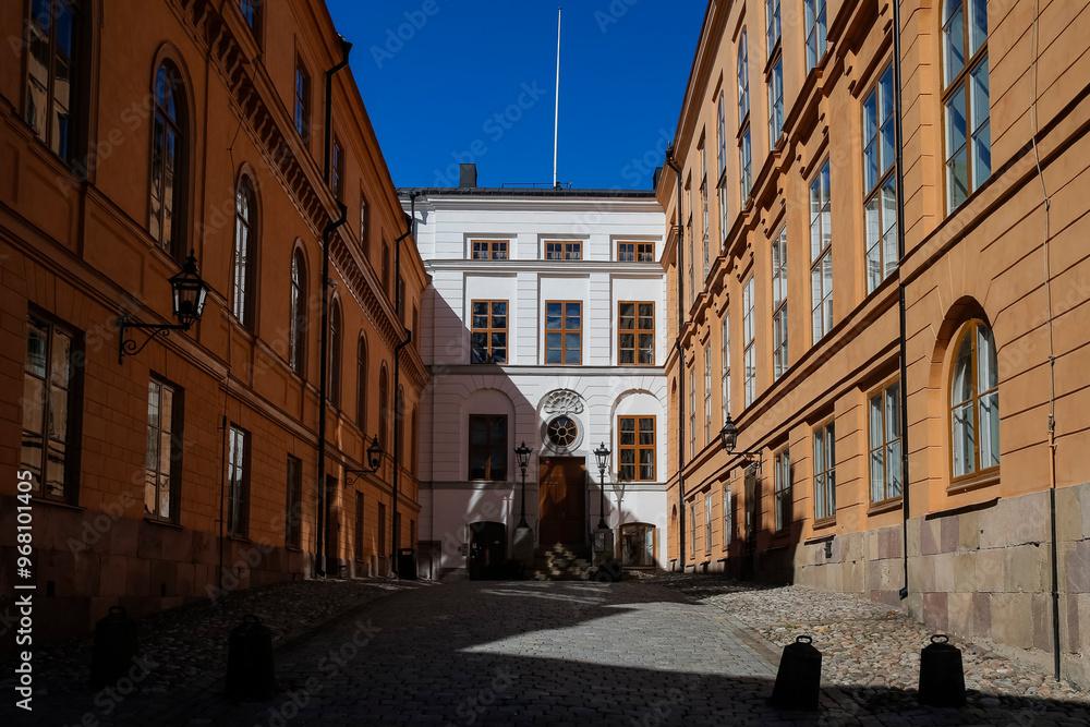 Fototapeta premium A peaceful cobblestone alleyway surrounded by warm-toned historic buildings captures the midday sunlight. The clear blue sky enhances the serene atmosphere, inviting exploration.