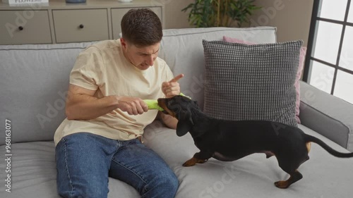 A young hispanic man in a living room admonishes his dachshund dog while sitting on a couch, indicating playful scolding indoors. photo