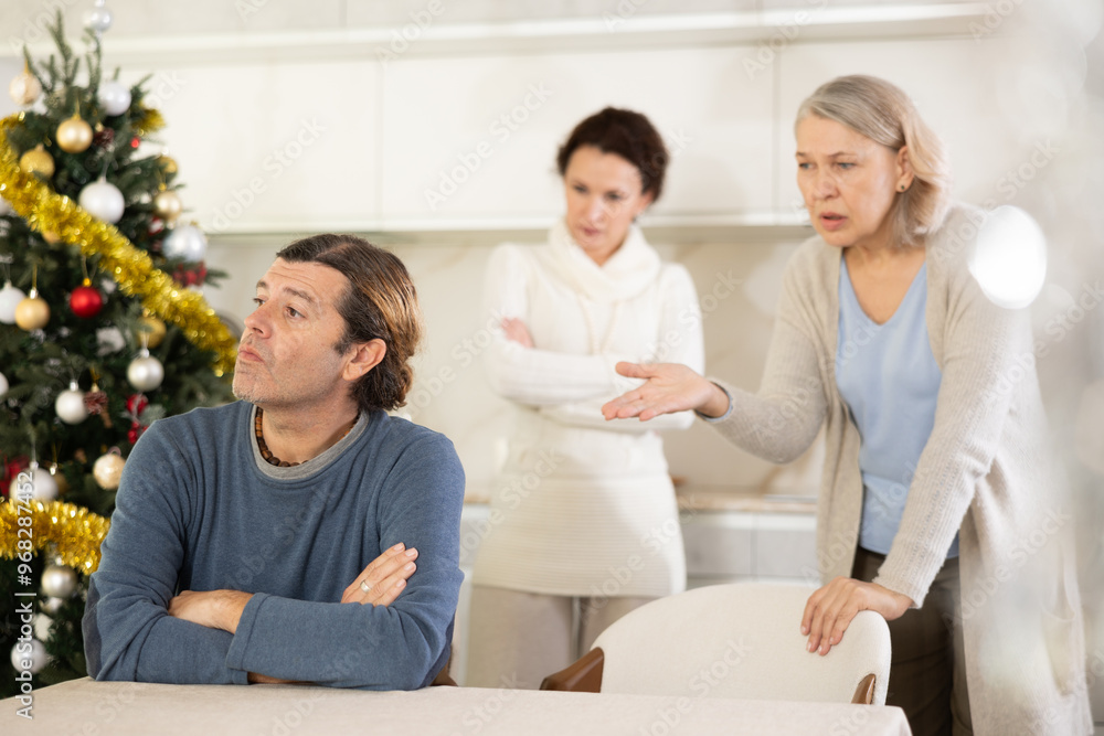 Obraz premium On Christmas Eve elderly mother and woman scold swear at man. Man is upset because of family conflict, dejectedly listens to his wife and mother scolding him. Complex intergenerational relationships
