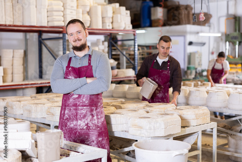 Portrait of smiling young bearded man, confident successful ceramicist, standing in stained maroon apron among racks with molds in pottery slip casting workshop photo