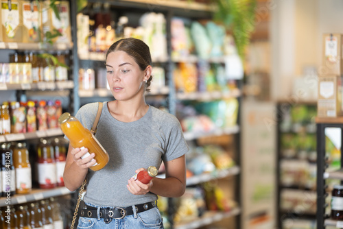 Interested young girl in casual gray t-shirt and jeans holding and examining organic juice bottles, pondering choices in natural food store