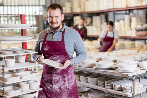 Enthusiastic young adult bearded ceramicist in maroon apron holding up freshly handcrafted plates, demonstrating high-quality standards maintained in ceramic studio.. photo