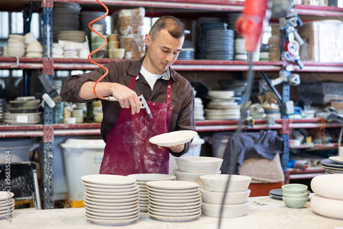 Experienced male ceramicist confidently using air blow gun to remove dust from ceramic dishes, actively working in artisan pottery studio photo