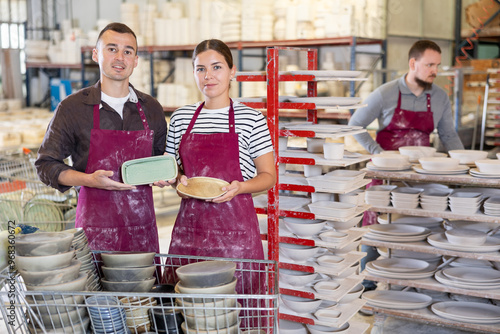 Smiling successful male and female artisans wearing maroon aprons, proudly displaying crafted ceramic plates in spacious workshop photo