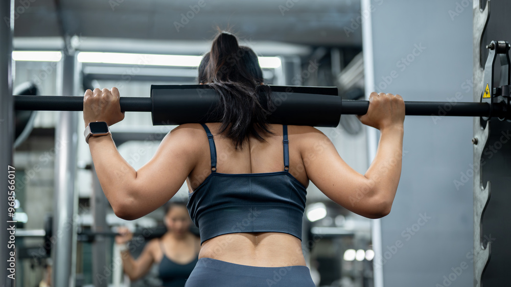 A back view of a sporty Asian woman in athletic wear placing a barbell on her shoulders. A back view of a sporty Asian woman in athletic wear placing a barbell on her shoulders.