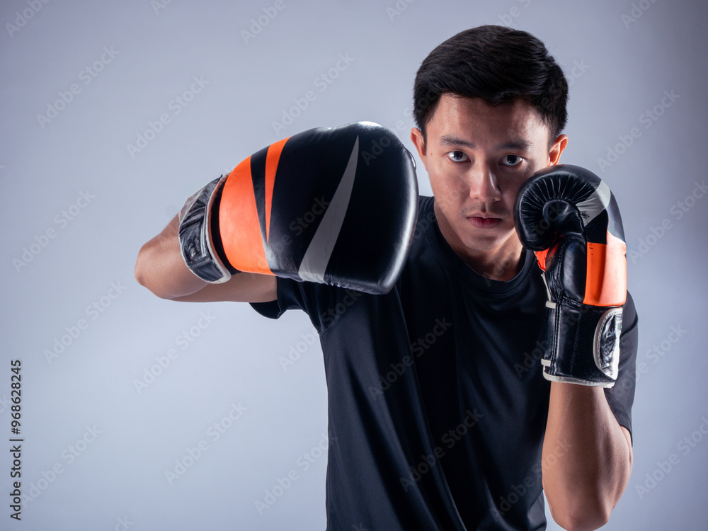 An Asian man in a serious boxing stance throws a punch while wearing black and orange boxing gloves. He is dressed in a black t-shirt and stands against a white background, ready for action An Asian man in a serious boxing stance throws a punch while wearing black and orange boxing gloves. He is dressed in a black t-shirt and stands against a white background, ready for action