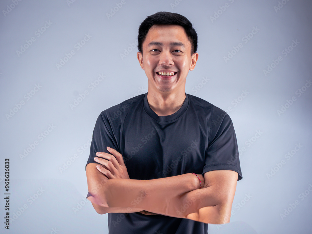An Asian man standing confidently with arms crossed, smiling while wearing a black t-shirt. He stands against a white background in a fitness studio, showcasing a positive and relaxed attitude An Asian man standing confidently with arms crossed, smiling while wearing a black t-shirt. He stands against a white background in a fitness studio, showcasing a positive and relaxed attitude