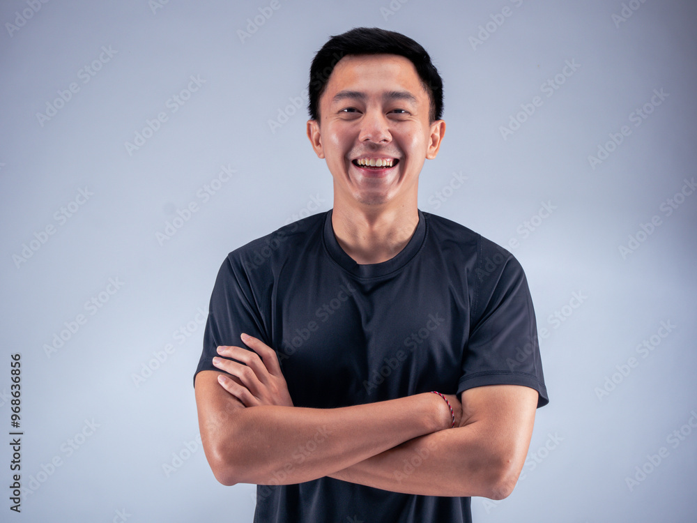 An Asian man standing confidently with arms crossed, smiling while wearing a black t-shirt. He stands against a white background in a fitness studio, showcasing a positive and relaxed attitude An Asian man standing confidently with arms crossed, smiling while wearing a black t-shirt. He stands against a white background in a fitness studio, showcasing a positive and relaxed attitude