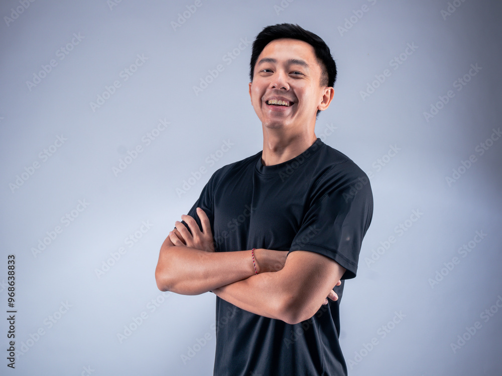 Fototapeta premium An Asian man standing confidently with arms crossed, smiling while wearing a black t-shirt. He stands against a white background in a fitness studio, showcasing a positive and relaxed attitude