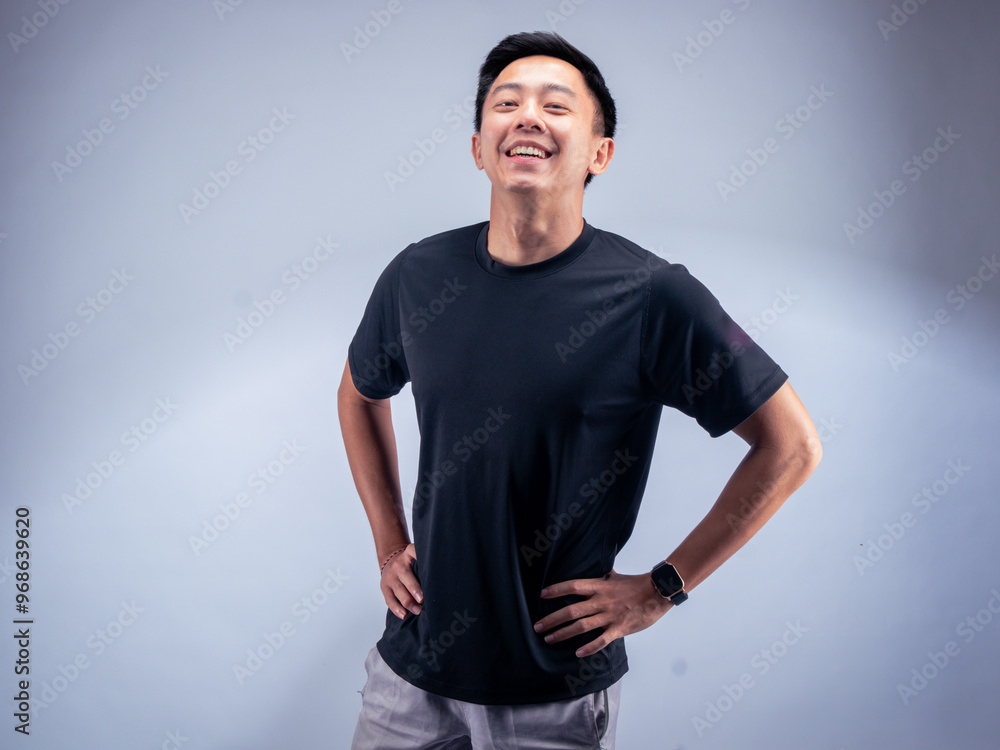 An Asian man stands with hands on hips, smiling confidently, wearing a black t-shirt and gray shorts. He poses against a white background in a fitness studio, showcasing a relaxed demeanor. An Asian man stands with hands on hips, smiling confidently, wearing a black t-shirt and gray shorts. He poses against a white background in a fitness studio, showcasing a relaxed demeanor.