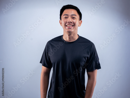 An Asian man smiling warmly while standing in a relaxed pose, wearing a black t-shirt. He is positioned against a white background in a fitness studio, conveying a sense of confidence