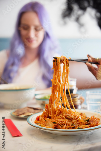 Cropped hand of young woman pulling noodles with chopsticks while friend sitting in background photo