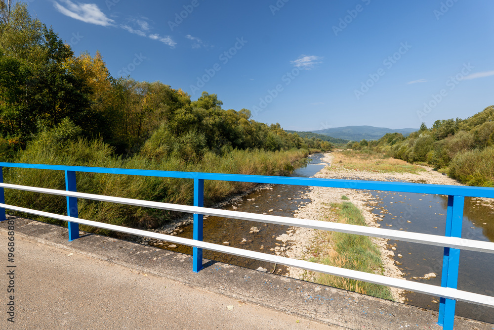 Beautiful mountain landscape on a sunny day.Natural coastline. A small amount of water in the river. Beskid Mountains. Bridge with blue and white balustrades.The Soła River. View from the