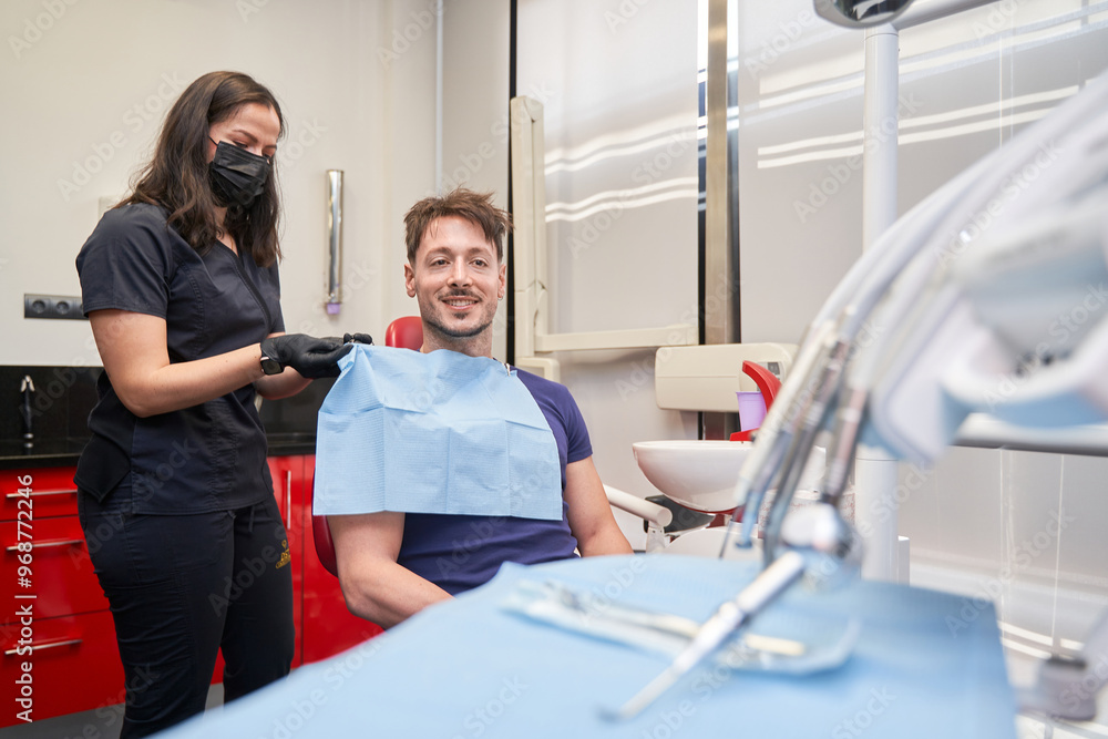 Obraz premium latina woman dentist putting a bib on a smiling man sitting in the chair to perform a procedure