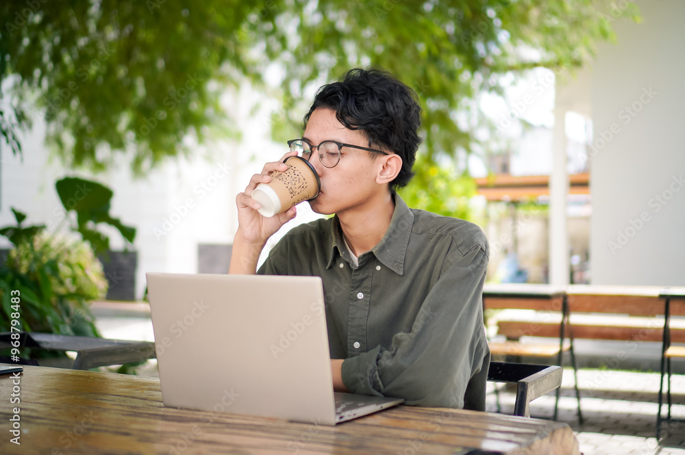 Young asian man thinking while looking to his laptop. Man working on his laptop at the outdoor cafe in the morning Young asian man thinking while looking to his laptop. Man working on his laptop at the outdoor cafe in the morning