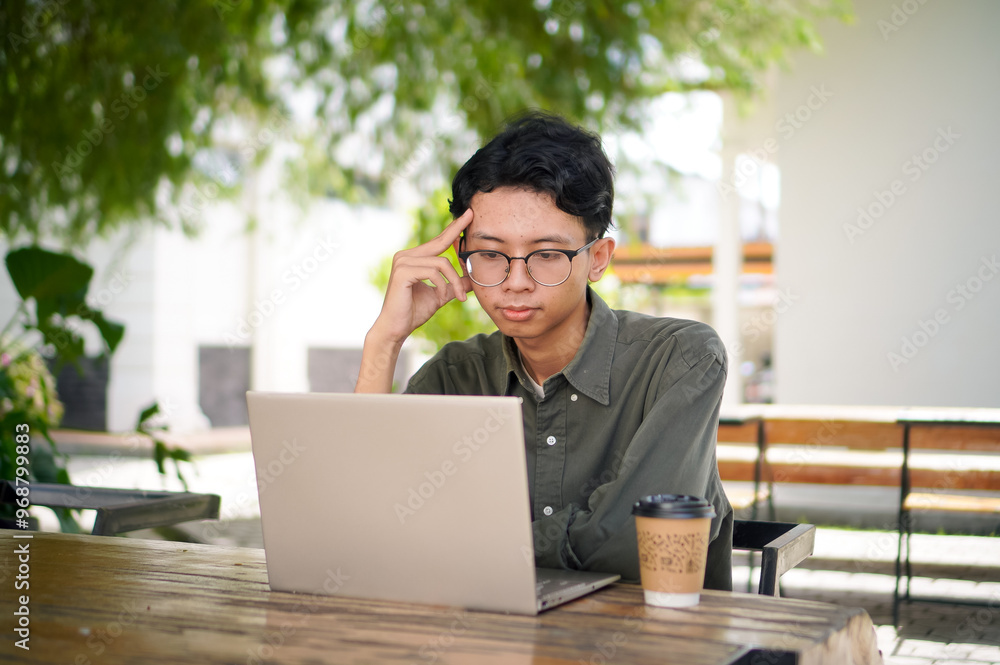 Young asian man thinking while looking to his laptop. Man working on his laptop at the outdoor cafe in the morning Young asian man thinking while looking to his laptop. Man working on his laptop at the outdoor cafe in the morning