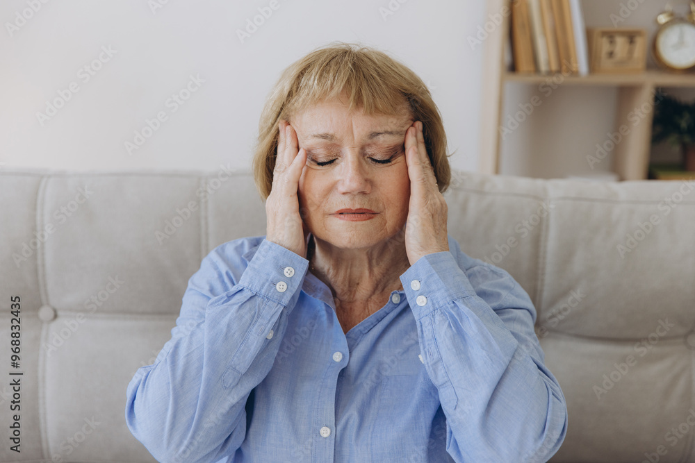© anatoliycherkas - Portrait of senior woman sitting on a sofa at home with a headache, feeling pain and with an expression of being unwell