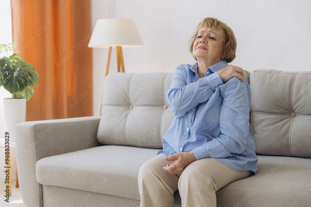 Senior woman sitting on sofa with shoulder pain. Elderly woman is enduring awful ache. Old woman suffering from shoulder injury, health problem concept Senior woman sitting on sofa with shoulder pain. Elderly woman is enduring awful ache. Old woman suffering from shoulder injury, health problem concept