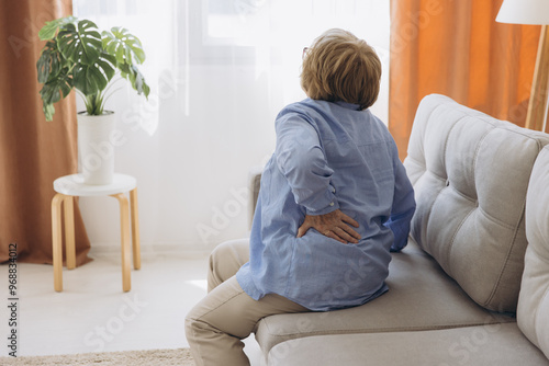 Unhappy old woman touching back, feeling pain, sitting on couch in living room, unhealthy middle aged senior female suffering from backache, spinal photo