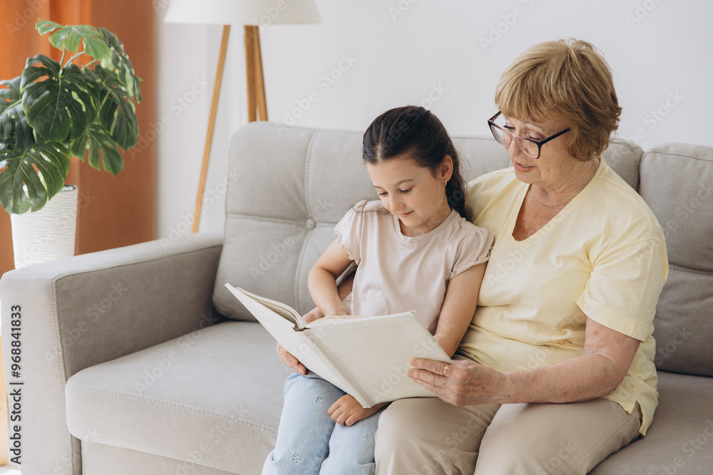 Happy loving Caucasian mature grandmother sit rest on sofa in living room read book with cute little granddaughter. Caring grandparent enjoy literature Happy loving Caucasian mature grandmother sit rest on sofa in living room read book with cute little granddaughter. Caring grandparent enjoy literature