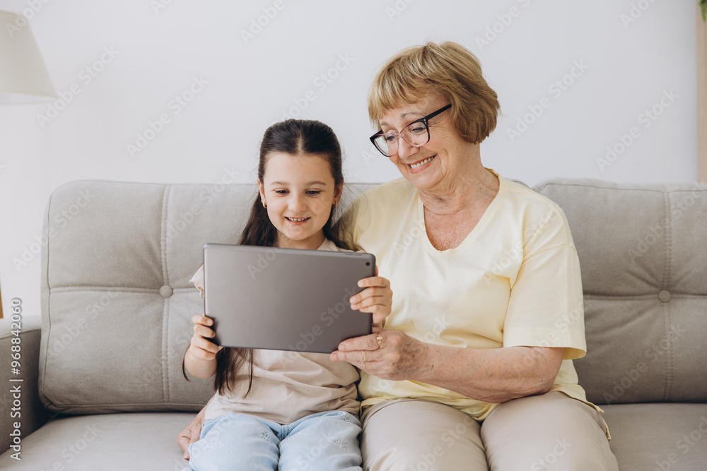 Having fun. Smiling retired woman nanny sit on cozy couch embrace little girl watch funny cartoon at web online using tablet. Bonding senior granny and small grandchild enjoy video clip on digital pad Having fun. Smiling retired woman nanny sit on cozy couch embrace little girl watch funny cartoon at web online using tablet. Bonding senior granny and small grandchild enjoy video clip on digital pad