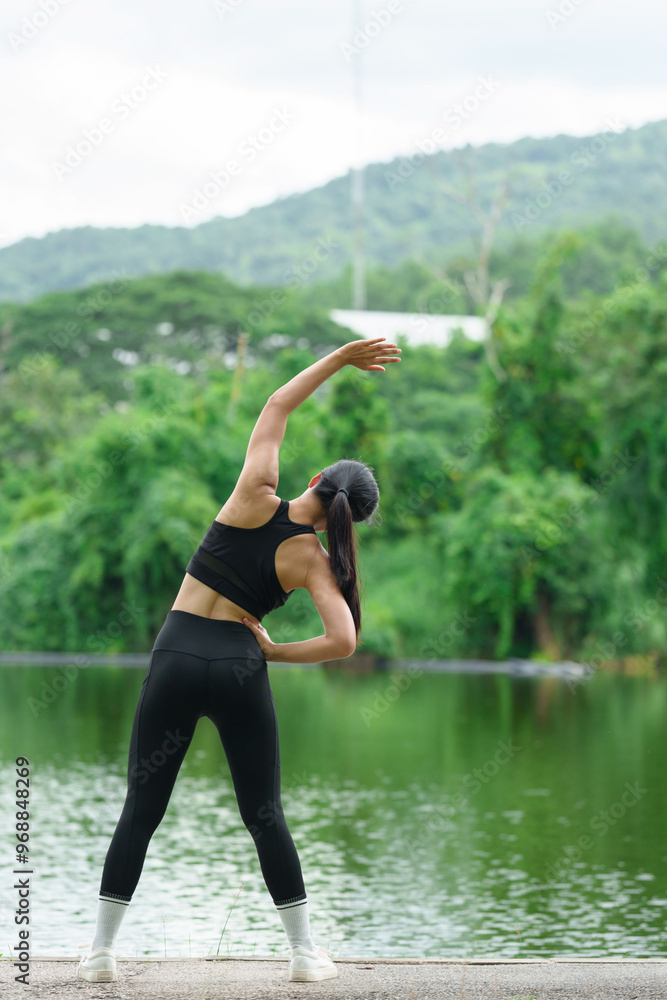 Fototapeta premium A young Asian woman wearing black exercise clothes, has a good figure, is strong, and is warming up. Stretch your legs before your run in a shady park. Exercise concept for health.