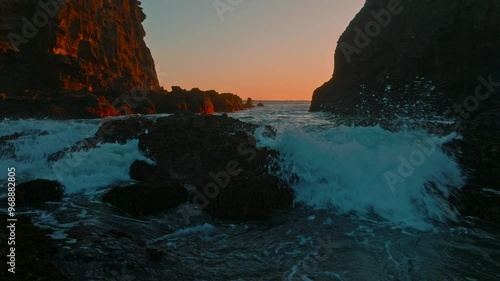 Ocean waves crashing through a cove over rocks and sand at sunset. Bethells Beach, Auckland, New Zealand.