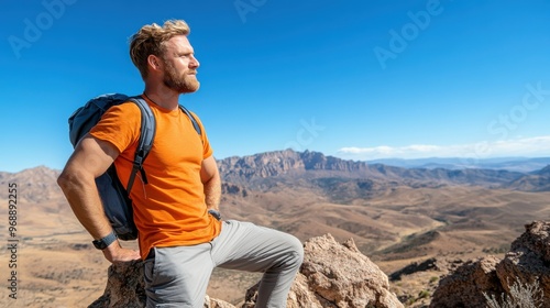 A man in an orange T-shirt, with a backpack, standing on a rocky terrain, looking at the vast mountainous landscape under a clear blue sky, symbolizing adventure and freedom. photo