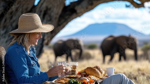 A woman with a hat enjoys a meal at a table in a scenic safari landscape, with elephants and mountains in the background, depicting an adventurous outdoor dining experience. photo