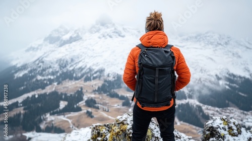 A hiker wearing an orange jacket and a backpack stands on a snowy mountain ridge, gazing thoughtfully out over a picturesque valley, encapsulating the essence of wilderness exploration. photo