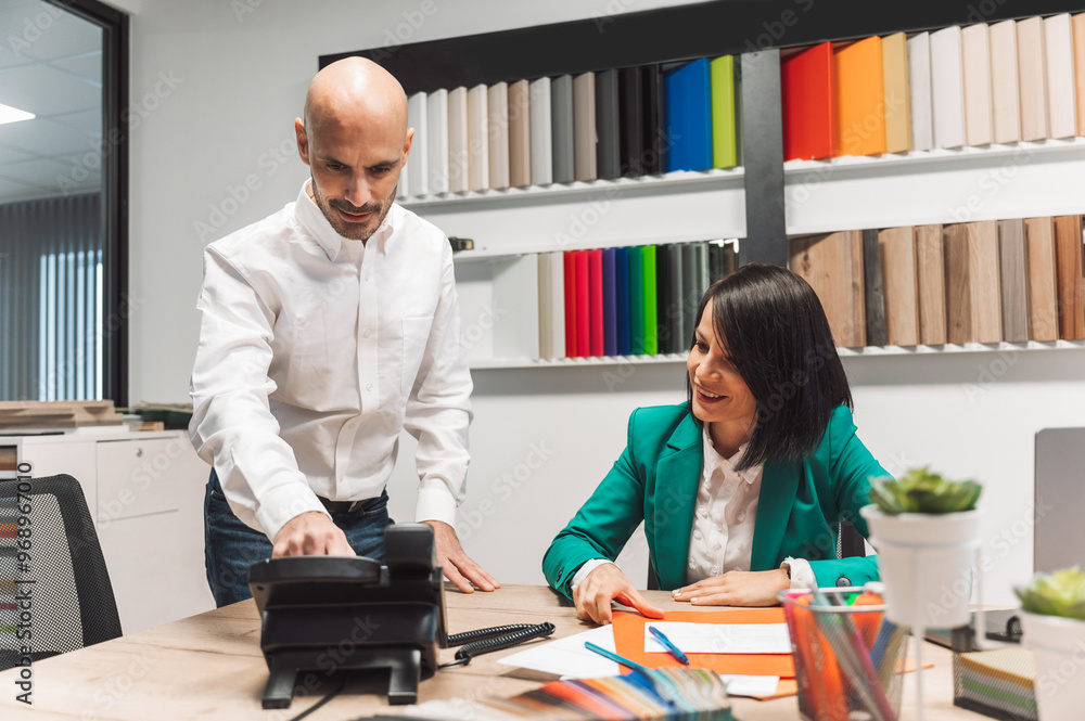 Obraz premium A professional meeting in an office setting. A man in a white shirt is showing something on a phone to a woman in a green blazer, who is smiling. The background features colorful materials and a moder