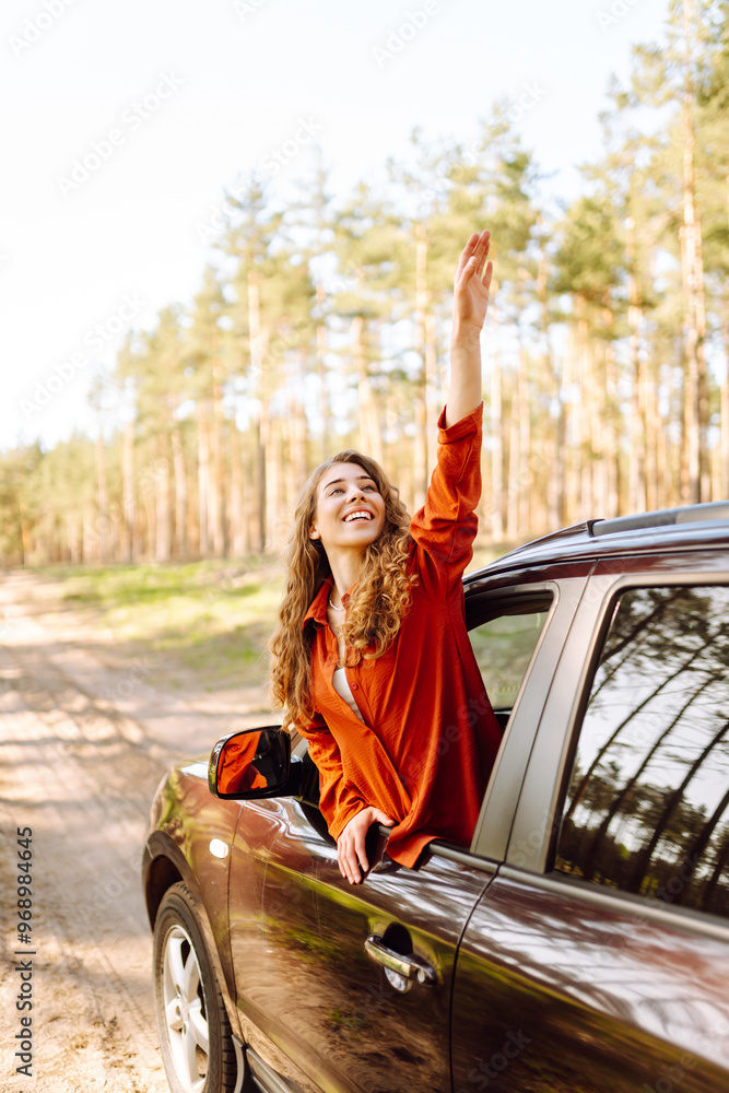 The young woman leans out of the car window, basking in the sunlight and enjoying the natural surroundings of a serene forest road. The concept of active lifestyle, travel, tourism.