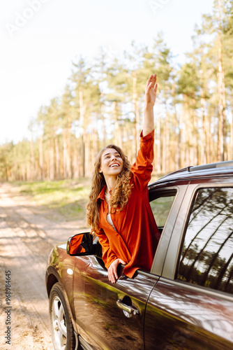 Obraz na płótnie The young woman leans out of the car window, basking in the sunlight and enjoying the natural surroundings of a serene forest road. The concept of active lifestyle, travel, tourism. w salonie