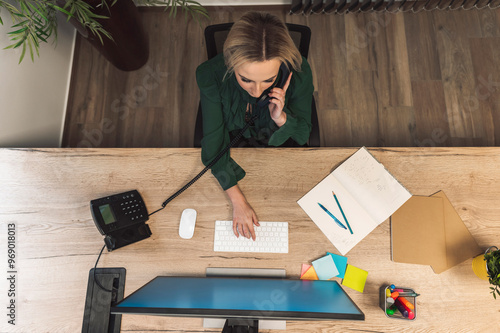 An overhead view of a woman working at a desk, talking on the phone while typing on a keyboard. The workspace includes a computer monitor, office supplies, and a notepad with a pen.