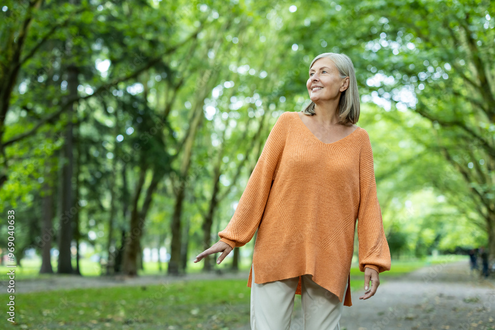 Mature woman walking through a serene park surrounded by vibrant greenery. She wearing an orange sweater, smiling, and expressing feelings of happiness, relaxation, and contentment enjoying nature. Mature woman walking through a serene park surrounded by vibrant greenery. She wearing an orange sweater, smiling, and expressing feelings of happiness, relaxation, and contentment enjoying nature.
