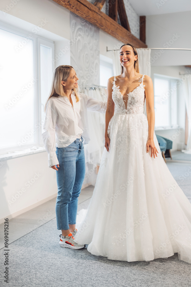 Naklejka premium A bridal designer fitting a wedding dress on a smiling bride in a bright, airy studio. The dress features intricate lace details and a flowing skirt, while the designer looks pleased with the fit.