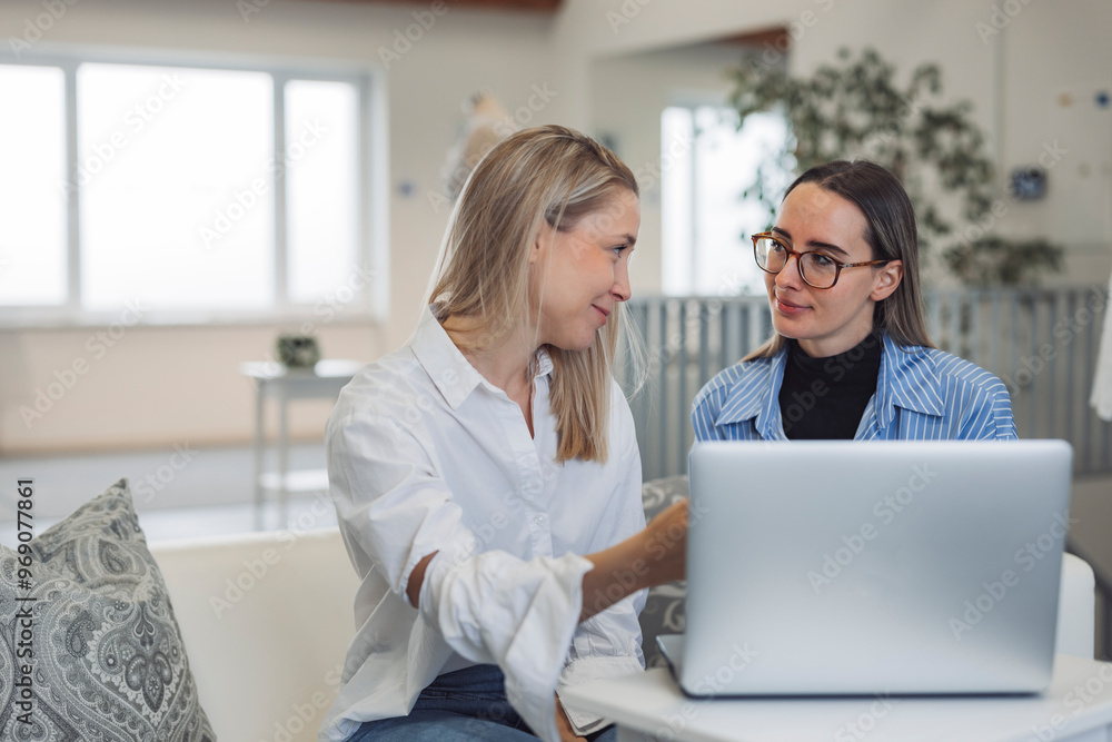 Two women smiling while looking at a laptop together in a bright, modern workspace. One woman has long blonde hair and is wearing a white shirt, while the other has long dark hair and glasses, dressed Two women smiling while looking at a laptop together in a bright, modern workspace. One woman has long blonde hair and is wearing a white shirt, while the other has long dark hair and glasses, dressed
