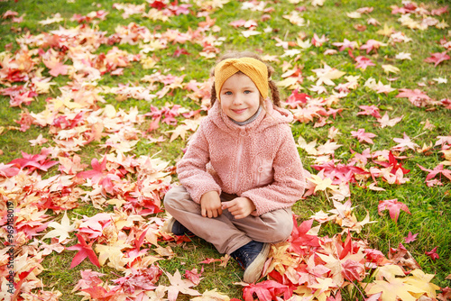 A smiling girl wearing a cozy sweater and headband sits cross-legged on a grassy area covered with colorful fallen leaves, enjoying the beauty of autumn.