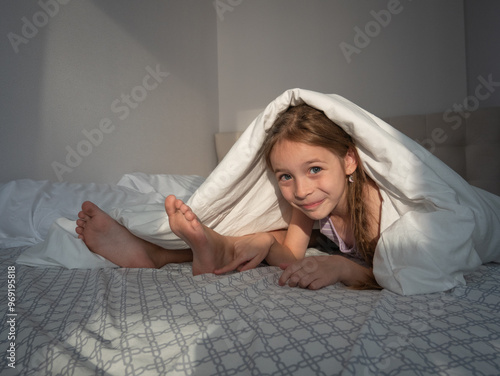 A little girl lies on her stomach on the bed, looks out from under the duvet, next to her sister's bare feet, smiles slyly, pulls her hands to her heels to tickle, laughs and looks into the frame photo