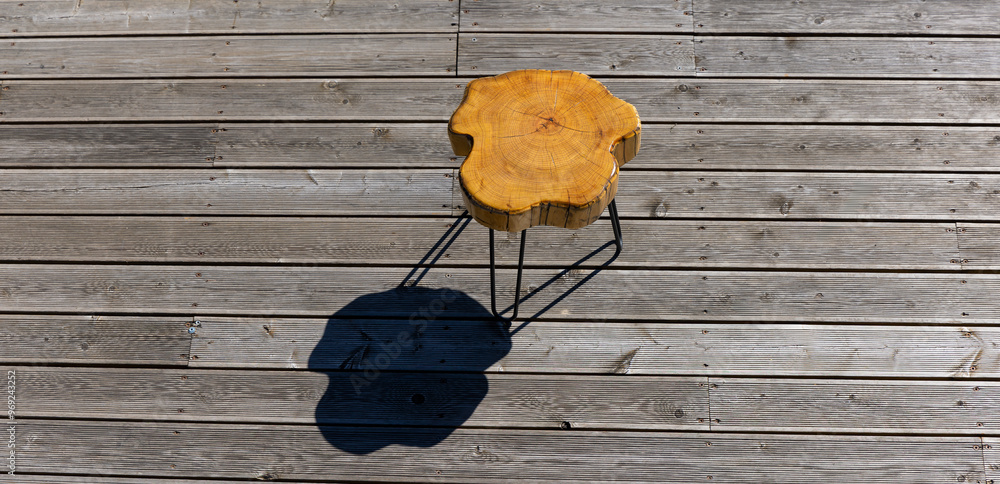 A coffe table made of a light wood trunk against the background of wooden terrace boards. The tree rings of the trunk are clearly visible. A shot of the table from above, taken on a sunny day.