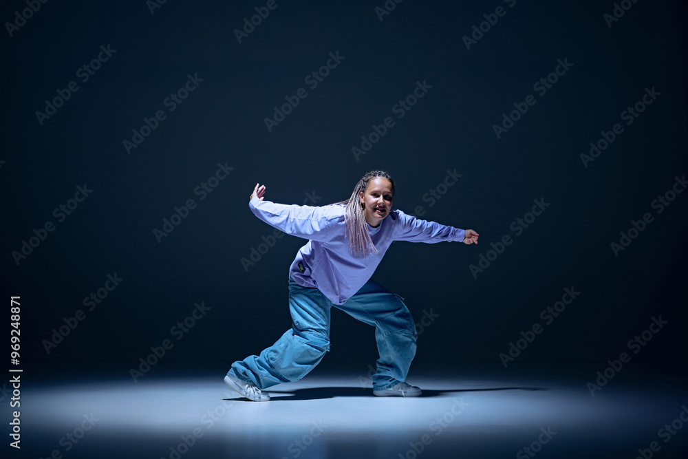 Dynamic image of young girl, passionate dancer in oversized clothes dancing hip hop with energy against navy blue studio background. Concept of freestyle dance, modernity, urban culture, youth Dynamic image of young girl, passionate dancer in oversized clothes dancing hip hop with energy against navy blue studio background. Concept of freestyle dance, modernity, urban culture, youth