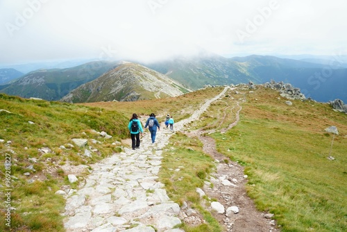 Hikers walk on a mountain trail with scenic views and a cloudy sky in the background.