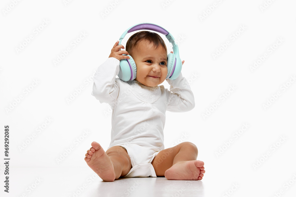 Happy little baby girl, child sitting in onesie listening to music, fairy tales on headphones isolated on white studio background. Concept of childhood, parenthood, care Happy little baby girl, child sitting in onesie listening to music, fairy tales on headphones isolated on white studio background. Concept of childhood, parenthood, care
