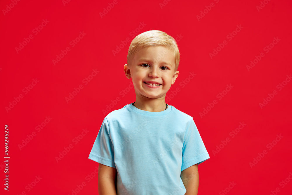 Portrait of little 4 years old boy with blonde hair, standing with cheerful smile, wearing blue t-shirt and posing against red studio background. Concept of childhood, emotions, fashion, lifestyle Portrait of little 4 years old boy with blonde hair, standing with cheerful smile, wearing blue t-shirt and posing against red studio background. Concept of childhood, emotions, fashion, lifestyle