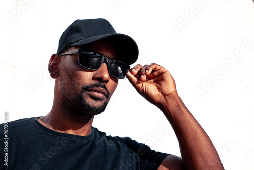 Afro black guy on the city center wearing black clothes cap an sun glasses modeling touching sunglasses with a white background.