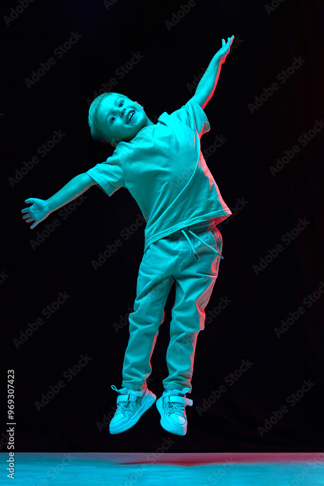 Full-length of happy little boy, 4 years old child in casual clothes cheerfully jumping, playing against black studio background in neon light. Concept of childhood, emotions, fashion, lifestyle Full-length of happy little boy, 4 years old child in casual clothes cheerfully jumping, playing against black studio background in neon light. Concept of childhood, emotions, fashion, lifestyle