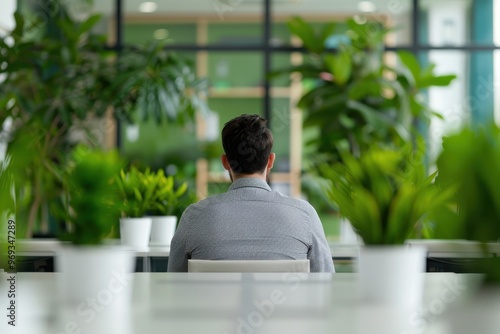 A man sits alone in a modern office space, surrounded by lush indoor plants, creating a peaceful and green workspace atmosphere, viewed from behind.