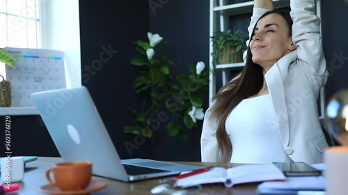 Relaxed woman leans back with her feet up on the desk, smiling satisfactorily in a modern office. The workspace includes a laptop, coffee cup, and notebook, creating a calm, casual work environment.